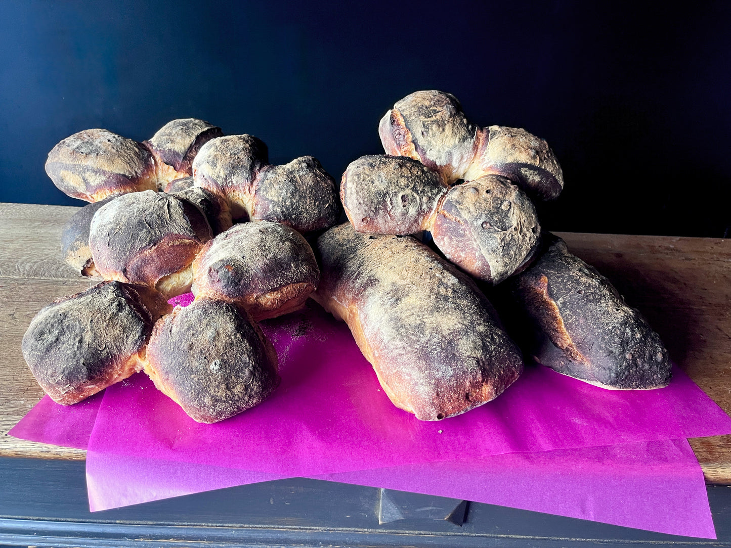 Auf einem rustikalen Holzregal liegen auf pinkem Papier insgesamt sechs Backwaren von bread.love: 4 Bürli und 2 Pfünderli. Die Bürli sind als vierteilige, rundlich zusammengewachsene Brötchen mit dunkler, bemehlter Kruste zu erkennen, während die Pfünderli länglich-oval geformt sind und eine kräftig gebackene, rustikale Oberfläche haben. Die Brote sind locker übereinander arrangiert und stehen vor einem dunklen Hintergrund.