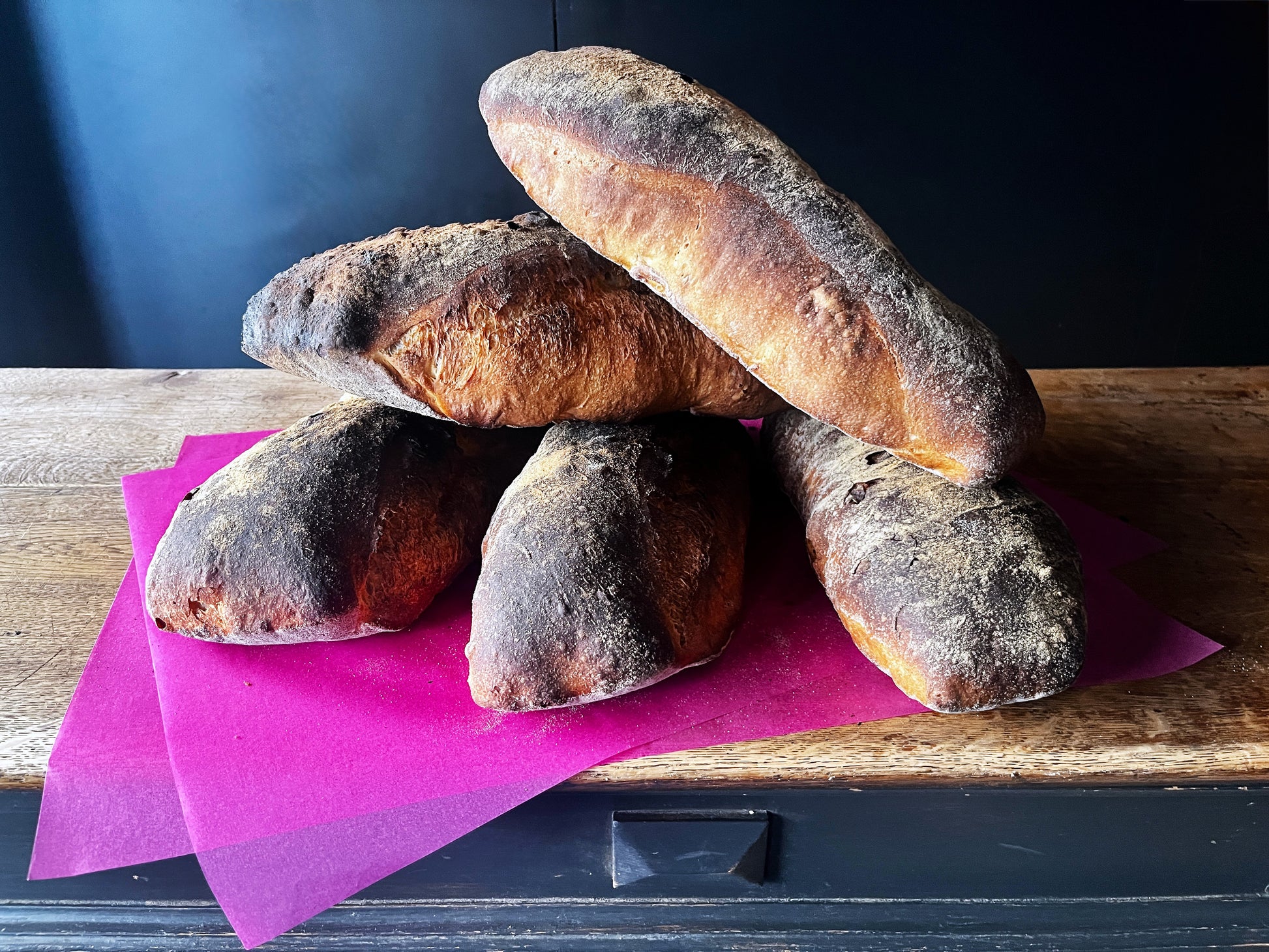 Fünf Pfünderli von bread.love, malerisch aufeinander gestapelt auf leuchtend pinkem Papier auf einem rustikalen Holzregal vor dunklem Hintergrund. Die Brote sind länglich-oval geformt mit abgerundeten Enden – ähnlich einem kleinen Batard. Ihre Kruste ist kräftig dunkel, mit mehligem Belag bestäubt und zeigt eine unregelmässige, handwerklich rustikale Oberfläche. An den Flanken schimmert die Kruste warm rötlich-golden durch, was auf einen intensiven Backprozess im Holzofen hindeutet.