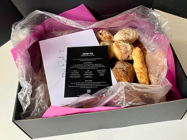 Geöffnete Versandbox von bread.love, ausgekleidet mit pinkem Seidenpapier und transparenter Schutzfolie. Darin liegen frisch gebackene Bio-Sauerteigbrote wie Bürli, Pfünderli und Yuzubängeli. Obenauf liegen eine persönliche, handgeschriebene Grußkarte sowie ein schwarzer Flyer mit der Aufschrift "HOW TO bread.love at home", der Anleitungen zum korrekten Aufbacken und Lagern der Brote enthält.