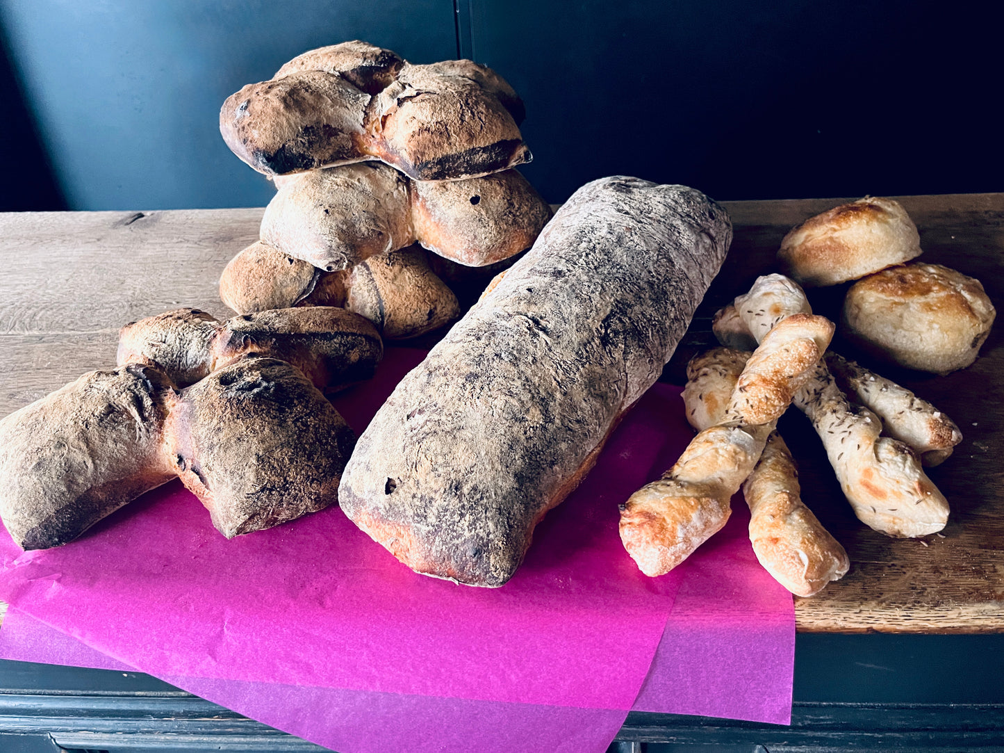 Eine Auswahl frisch gebackener Brote und Kleingebäcke von bread.love auf einem rustikalen Holzregal vor dunklem Hintergrund, arrangiert auf leuchtend pinkem Papier. Links und im Hintergrund sind mehrere Bürli gestapelt – rundlich-ovale, dunkle Brötchen mit grober, knuspriger Kruste und sichtbaren dunklen Einschlüssen. In der Mitte liegt ein längliches, zylindrisches Pfünderli mit kräftig dunkler, mehlig bestäubter Kruste. Rechts vorne liegen mehrere Yuzubängeli – dünne, gedrehte Brotstangen mit goldbrauner 